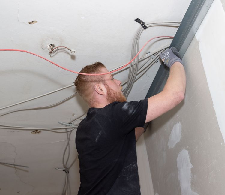 Junger Elektriker mit schwarzem T-Shirt verzieht Kabel mittels Aluminium-Profilschiene unter einer Zimmerdecke.