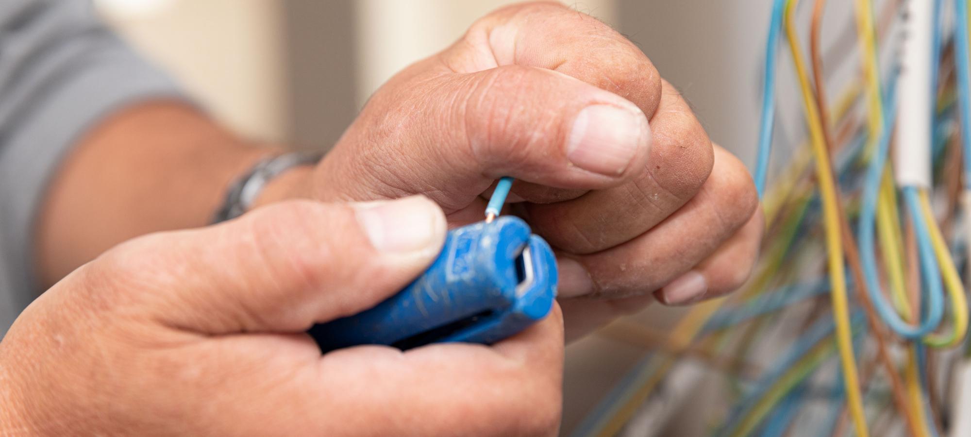 Bildausschnitt mit zwei Händen eines Elektromeisters beim abisolieren eines blauen Kabels mittels Abisolierzange.