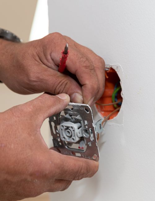 Bildausschnitt zweier Hände eines Elektromeisters bei der Feinmontage einer Steckdose an einer weißen Wand.