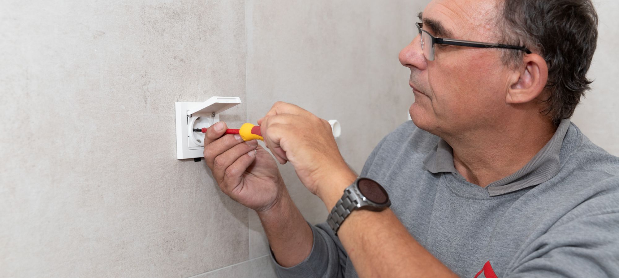 Elektriker mit Schraubendreher schließt eine weiße Steckdose an einer grau gefliesten Wand an